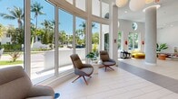 Located at 2500 SW 22nd Ter in Fort Lauderdale, Florida, this single-floor space is a striking example of contemporary design that seamlessly blends artistry with functionality. The interior is immediately distinguished by its soaring double-height ceilings, dramatic floor-to-ceiling windows, and circular porthole accents that flood the space with an abundance of natural light and frame lush views of surrounding palm trees and tropical landscaping.

The main living and lounge area serves as the heart of the space, featuring light-toned large-format tile flooring, a curated mix of modern seating including swivel accent chairs and a curved lounge sofa, and a bold, multicolored vertical striped wall mural that adds a dynamic artistic focal point. Globe pendant chandeliers and wall-mounted red sconces provide layered, stylish lighting throughout.

A glass-partitioned office area offers a dedicated workspace with a refined, professional atmosphere, while maintaining visual connectivity to the main floor through its transparent walls. The hallway provides a clean transitional corridor connecting the primary areas to the ADA-accessible bathroom, which is finished with large-format dark tile flooring and walls, a wall-mounted sink, and a clean, utilitarian aesthetic.

Overall, the space exudes a bright, modern, and energetic ambiance — an inviting environment defined by artistic expression, ample natural light, and thoughtful design.