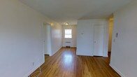 This single-floor space presents a clean, straightforward layout designed for comfortable everyday living. The main living area is highlighted by rich, warm-toned hardwood floors that run throughout, adding a timeless character to the interior. Bright white walls and white trim create a crisp, neutral backdrop that allows natural light to reflect and brighten the space. A window positioned at the far end of the room invites in natural daylight, contributing to an open and airy feel. A baseboard radiator beneath the window provides efficient heating during cooler months. The room features multiple doorways along its perimeter, offering seamless access to adjoining spaces and suggesting a well-connected floor plan. A flush-mount ceiling light fixture provides overhead illumination, ensuring the space remains well-lit throughout the day and evening. The overall ambiance is clean and understated, with a move-in-ready appeal. The combination of classic hardwood flooring, neutral tones, and functional layout makes this a versatile space adaptable to a variety of furnishing styles and personal preferences.
