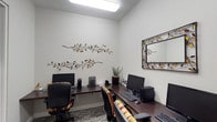 This single-floor business center offers a functional and thoughtfully designed workspace equipped with multiple computer workstations, each featuring a desktop computer, keyboard, and mouse setup arranged along dark wood-finish desks. The workstations are complemented by ergonomic office chairs with upholstered striped cushioning, ensuring comfort during use. The space is finished with clean white walls that create a bright, open feel, further enhanced by recessed and overhead fluorescent lighting. Decorative metal branch-and-leaf wall sculptures add an artistic touch, lending warmth and character to the otherwise professional environment. A framed mirror with a matching decorative metal border is mounted on the wall, contributing to the cohesive aesthetic. A potted plant provides a subtle natural accent. The flooring features a patterned low-pile carpet that adds texture underfoot. The overall ambiance is clean, orderly, and welcoming, making this space well-suited for tasks such as printing, browsing, or remote work in a shared setting.