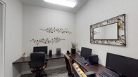 This single-floor business center offers a functional and thoughtfully designed workspace equipped with multiple computer workstations, each featuring a desktop computer, keyboard, and mouse setup arranged along dark wood-finish desks. The workstations are complemented by ergonomic office chairs with upholstered striped cushioning, ensuring comfort during use. The space is finished with clean white walls that create a bright, open feel, further enhanced by recessed and overhead fluorescent lighting. Decorative metal branch-and-leaf wall sculptures add an artistic touch, lending warmth and character to the otherwise professional environment. A framed mirror with a matching decorative metal border is mounted on the wall, contributing to the cohesive aesthetic. A potted plant provides a subtle natural accent. The flooring features a patterned low-pile carpet that adds texture underfoot. The overall ambiance is clean, orderly, and welcoming, making this space well-suited for tasks such as printing, browsing, or remote work in a shared setting.