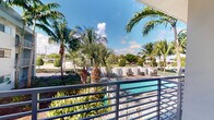 This single-story space offers a standout outdoor feature: a private balcony enclosed by sleek horizontal metal railings, providing direct views of a shared swimming pool below. The pool area is well-appointed with lounge seating and is framed by an abundance of mature tropical palm trees, creating a lush, resort-like atmosphere. The surrounding landscape is verdant and well-maintained, with layered greenery adding depth and privacy to the outdoor setting. The balcony benefits from generous natural light and open sky exposure, making it a practical and inviting space for outdoor relaxation. The tropical setting and bright, sunny skies reinforce a warm-climate ambiance throughout. The remaining interior images were too underexposed to provide reliable detail, so interior features of the space are not described here. Overall, the outdoor balcony and pool-facing orientation are the defining characteristics of this property, lending it a distinctly tropical, leisure-oriented character well-suited to a warm-weather lifestyle.