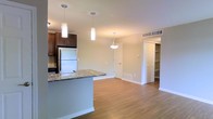 This single-floor residence showcases an open-concept layout that seamlessly connects the kitchen and living areas. The kitchen is equipped with dark espresso cabinetry, a stainless steel refrigerator, and a granite-topped island that doubles as a breakfast bar, illuminated by two sleek cylindrical pendant lights. The flooring throughout is warm hardwood-style, adding continuity and a sense of spaciousness across the main living area. A third decorative pendant light fixture anchors the dining or living zone, enhancing the open feel of the space. Neutral beige walls provide a clean, versatile backdrop throughout. Ventilation is well-managed with ceiling-mounted HVAC vents positioned strategically. A closet with built-in shelving is visible near the far wall, offering organized storage solutions. White panel doors and bright white baseboards complement the overall clean aesthetic. The layout appears efficient and functional, with clear separation between living and storage zones while maintaining an airy, connected atmosphere suitable for everyday living.