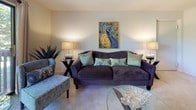 Located at 710 Snowberry Court in Baltimore, MD, this single-floor residence offers a well-organized layout with distinct living, dining, sleeping, and kitchen areas. The living room is a welcoming space anchored by a plush sofa accented with decorative pillows, flanked by matching table lamps, and adorned with a vibrant peacock-themed artwork that serves as a focal point on the neutral-toned walls. Carpeted flooring runs throughout, adding warmth to the interior. The dining area features a glass-top round table surrounded by elegantly upholstered chairs with decorative nail-head trim, complemented by a substantial dark wood hutch offering ample storage. A decorative sunburst mirror adds a refined touch to the dining space. The kitchen connects to the main living area via a pass-through breakfast bar fitted with turquoise barstools, providing a casual dining option. Light cabinetry and under-cabinet lighting keep the kitchen bright and functional. The primary bedroom is generously proportioned, featuring a tufted headboard, coordinated bedside tables, and soft ambient lighting, creating a serene retreat. A secondary bedroom includes a window with views of lush greenery. The bathroom is clean and efficiently designed with a tub-shower combination, vanity with storage, and classic hexagonal tile flooring.