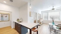 This thoughtfully designed 2-bedroom, 1-bathroom residence features a clean, modern aesthetic throughout, with light hardwood-style flooring and crisp white walls creating a bright, cohesive feel across all rooms. The open-concept kitchen and living area serves as the heart of the home, anchored by a peninsula-style island with a white quartz countertop and bar seating — ideal for casual dining or entertaining. Dark cabinetry contrasts elegantly with the light countertops, while a suite of stainless steel appliances, including a refrigerator, electric range, microwave, and dishwasher, equips the space for everyday cooking. A glass pendant light adds a refined touch overhead. The living area offers generous wall space and natural light from nearby windows. Both bedrooms are comfortably sized, each featuring windows with blinds that allow for natural light control, and ceiling fixtures. The bathroom includes a floating vanity with dark cabinetry, open built-in shelving for storage, and a separate tub-shower combination. A dedicated laundry room houses a full-size washer and dryer. The unit also includes a closet for additional storage. Overall, the space presents a clean, contemporary design with functional, well-appointed rooms throughout.