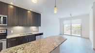 This single-floor unit showcases a contemporary open-concept design with light wood-tone flooring throughout. The kitchen is a standout feature, equipped with dark espresso cabinetry, a tile backsplash, stainless steel appliances including a refrigerator, dishwasher, and microwave, and a generous granite-topped island with pendant lighting overhead. The island doubles as a breakfast bar, seamlessly connecting the kitchen to the living area, which benefits from natural light streaming through a large glass balcony door overlooking a green outdoor setting.

The bedroom is a well-proportioned, bright space with a ceiling fan, a window with a roller shade, and light-toned flooring. It connects directly to a spacious walk-in closet outfitted with built-in shelving and hanging rods. The bathroom features large-format marble-look white tile flooring, a dark-toned double vanity with an illuminated mirror, and a glass-enclosed shower. In-unit laundry is conveniently tucked behind louvered barn-style doors near the kitchen. Clean white walls, recessed lighting, and a neutral palette contribute to the overall bright, modern ambiance throughout the space.