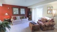 This two-floor home offers a thoughtfully designed interior with a welcoming atmosphere. The living room serves as a central gathering space, highlighted by a rich, deep-red accent wall that anchors the room and provides a striking visual contrast against the neutral cream-toned walls and light carpet flooring. Two large botanical framed prints adorn the accent wall, adding a classic, decorative touch.

The seating arrangement features a generously sized striped sofa complemented by a floral-upholstered loveseat and matching ottoman, creating a cohesive and comfortable layout. A tall wicker storage shelf tucked into the corner adds both functionality and warmth to the room's design.

Natural light flows generously into the living area through a large sliding glass door dressed with sheer white curtains, offering a view and access to an outdoor area. The soft carpeted flooring enhances the room's cozy ambiance. The overall design aesthetic balances traditional furnishings with warm, saturated color choices, resulting in a space that feels both elegant and livable across its two levels.