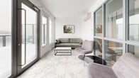 Located at 150 Rue King in Montréal, this mezzanine-level lounge is a compact yet well-appointed space that blends modern design with a warm, inviting atmosphere. The room is defined by its striking marble-effect porcelain tile flooring, which runs throughout and lends an air of elegance and visual spaciousness to the interior.

Large windows along the exterior wall flood the space with natural light, while a set of full-height French doors provides direct access to an outdoor balcony, offering views of the surrounding urban environment. A wall-mounted mini-split climate control unit ensures year-round comfort.

The layout is thoughtfully arranged into two distinct seating areas. One area features a sage green upholstered sofa paired with a minimalist black-framed coffee table, anchored by botanical wall art that reinforces the space's nature-inspired aesthetic. The second area offers a pair of wooden sling chairs with geometric-patterned upholstery, arranged around a wire-frame side table beside a towering Bird of Paradise plant, which adds a lush, organic element to the crisp white surroundings.

Bright white walls and a clean white ceiling enhance the sense of openness, while the carefully curated furnishings create a cohesive, contemporary feel. This mezzanine lounge serves as a refined and functional communal or transitional space within the building.
