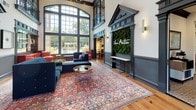 This distinguished space makes an immediate impression with its dramatic double-height ceilings and an abundance of natural light pouring through expansive arched and multi-pane windows. The main gathering area commands attention with its grand proportions, showcasing dark-painted classical columns, ornate pediment doorways, and an upper-level interior balcony with wrought-iron railings that overlook the main floor below. Rich hardwood flooring runs throughout, anchored by a large, vibrant Persian-style area rug that adds warmth and color to the refined setting. Seating areas are thoughtfully appointed with tufted navy velvet sofas, classic upholstered armchairs, and gold-accented side tables, all contributing to an atmosphere of sophisticated elegance. A striking living room wing features a stately black fireplace flanked by symmetrical windows, decorative wainscoting, and bold dark trim that contrasts beautifully against crisp white walls. An adjoining room houses a grand piano beneath tall windows dressed with floral valances and flowing curtains, creating an intimate and cultured retreat. A living moss wall bearing a branded insignia adds a contemporary organic touch near the entrance. The space masterfully balances classical architectural heritage with curated interior design, resulting in an environment that is both impressive and inviting.