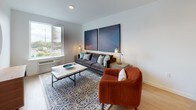 This well-designed single-floor residence showcases a thoughtful open-concept layout that seamlessly connects the kitchen, dining, and living areas. The contemporary kitchen is a standout feature, equipped with rich dark wood cabinetry, white quartz countertops, stainless steel appliances including a gas range, microwave, and full-size refrigerator, and a large center island with seating for multiple bar stools — ideal for both meal prep and casual dining. Light wood-style flooring runs throughout the main living areas, adding warmth to the clean, modern aesthetic. The living room offers a comfortable, stylish setting with ample natural light streaming through a large window that frames outdoor views of the surrounding urban landscape, complemented by recessed lighting and generous wall space for art. The bedroom continues the bright, airy feel of the home, featuring large windows with expansive city views, plush carpeting, and a well-organized closet. The full bathroom is finished with the same warm wood cabinetry found throughout the home, paired with light flooring, a combined tub and shower, and a single vanity with a bright mirror. In-unit laundry is also present, adding everyday convenience to this refined and functional living space.