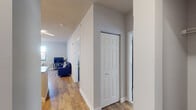 This well-designed single-floor residence offers a thoughtful layout that balances comfort and functionality. The open-concept kitchen serves as a central hub, featuring granite countertops on a spacious island with a built-in sink, modern stainless steel appliances including a refrigerator, range, and microwave, and crisp white cabinetry. Stylish angular ceiling light fixtures add a contemporary touch, while bar seating at the island makes for convenient casual dining. The kitchen flows seamlessly into the living area, which is finished with warm hardwood-style flooring and receives natural light through a large window overlooking the surrounding urban landscape. The living space comfortably accommodates a full seating arrangement centered around a flat-screen television. The bedroom is carpeted for added comfort and features a window that brings in natural light, offering views of the cityscape. It provides ample room for a full bedroom suite with generous wall space for storage furnishings. The bathroom is finished to a high standard, showcasing a granite-topped vanity with a white cabinet base, an LED-backlit mirror, and a full shower-tub combination with clean white tile surround. A hallway with hardwood-style flooring and a built-in closet connects the rooms, completing this efficiently planned and attractively finished space.