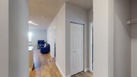This well-designed single-floor residence offers a thoughtful layout that balances comfort and functionality. The open-concept kitchen serves as a central hub, featuring granite countertops on a spacious island with a built-in sink, modern stainless steel appliances including a refrigerator, range, and microwave, and crisp white cabinetry. Stylish angular ceiling light fixtures add a contemporary touch, while bar seating at the island makes for convenient casual dining. The kitchen flows seamlessly into the living area, which is finished with warm hardwood-style flooring and receives natural light through a large window overlooking the surrounding urban landscape. The living space comfortably accommodates a full seating arrangement centered around a flat-screen television. The bedroom is carpeted for added comfort and features a window that brings in natural light, offering views of the cityscape. It provides ample room for a full bedroom suite with generous wall space for storage furnishings. The bathroom is finished to a high standard, showcasing a granite-topped vanity with a white cabinet base, an LED-backlit mirror, and a full shower-tub combination with clean white tile surround. A hallway with hardwood-style flooring and a built-in closet connects the rooms, completing this efficiently planned and attractively finished space.