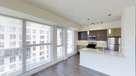 This single-floor space showcases a bright, open-concept layout that seamlessly integrates the kitchen and living area. A wall of large floor-to-ceiling windows floods the interior with natural light while offering views of the surrounding cityscape, creating an airy and expansive atmosphere. The wide-plank hardwood flooring runs throughout, adding warmth and continuity to the space.

The kitchen is a standout feature, designed with dark espresso cabinetry that contrasts elegantly against the crisp white quartz waterfall island, which also functions as a breakfast bar. Stainless steel appliances, including a gas range and built-in microwave, are integrated seamlessly into the cabinetry. Overhead, a combination of pendant lighting and a track lighting system provides both ambient and task illumination. Light taupe lower cabinetry along the back wall balances the darker upper units, contributing to a layered, sophisticated palette. The open layout allows the kitchen to flow naturally into the living area, making the overall space feel cohesive, functional, and well-suited for modern everyday living.