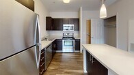 Located at 150 Exchange St in Malden, Massachusetts, this single-floor residential unit offers a clean, contemporary layout designed for comfortable everyday living. The kitchen is a standout feature, outfitted with dark espresso cabinetry, light countertops, and a full suite of stainless steel appliances including a refrigerator, range, built-in microwave, and dishwasher. Wood-look flooring grounds the kitchen space and extends into the adjoining area, while pendant lighting above the island adds a functional yet stylish touch. The open living area is generously proportioned, with neutral-toned walls, plush carpeting, and access to the kitchen, creating a seamless flow between spaces. Large windows in the bedroom allow abundant natural light to fill the room, offering views of the surrounding neighborhood. The bedroom also features a closet with panel doors, adding practical storage. The bathroom is finished with a dark vanity cabinet, white countertop, tile flooring, and a full soaking tub with shower enclosure. Throughout the unit, the design aesthetic is modern and understated, with a neutral color palette, clean lines, and quality finishes that provide a comfortable and move-in ready environment.