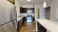 Located at 150 Exchange St in Malden, Massachusetts, this single-floor residential unit offers a clean, contemporary layout designed for comfortable everyday living. The kitchen is a standout feature, outfitted with dark espresso cabinetry, light countertops, and a full suite of stainless steel appliances including a refrigerator, range, built-in microwave, and dishwasher. Wood-look flooring grounds the kitchen space and extends into the adjoining area, while pendant lighting above the island adds a functional yet stylish touch. The open living area is generously proportioned, with neutral-toned walls, plush carpeting, and access to the kitchen, creating a seamless flow between spaces. Large windows in the bedroom allow abundant natural light to fill the room, offering views of the surrounding neighborhood. The bedroom also features a closet with panel doors, adding practical storage. The bathroom is finished with a dark vanity cabinet, white countertop, tile flooring, and a full soaking tub with shower enclosure. Throughout the unit, the design aesthetic is modern and understated, with a neutral color palette, clean lines, and quality finishes that provide a comfortable and move-in ready environment.