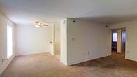 This single-floor space presents a clean, functional layout with a neutral aesthetic throughout. The main living area is finished with wall-to-wall carpeting in a warm beige tone, providing a soft and comfortable underfoot feel. Smooth, cream-colored walls offer a blank canvas well-suited for personalization.

A ceiling fan with an integrated light fixture is installed overhead, contributing to both air circulation and ambient lighting in the main living zone. HVAC vents are positioned along the walls and ceiling, ensuring climate control throughout the space. A thermostat is visibly mounted on the wall, offering convenient temperature management.

An open passage connects the main living area to an adjoining room or hallway, suggesting a straightforward and accessible floor plan. The partial wall dividing these zones adds a degree of definition without fully closing off the space, maintaining an open feel. Additional electrical outlets are distributed along the baseboards, providing practical utility throughout.

Overall, the space is characterized by its simplicity, neutral finishes, and practical layout, making it a versatile and move-in-ready environment.