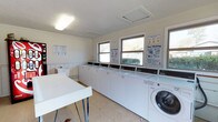 This single-floor communal laundry facility is designed for efficiency and ease of use. The room is bright and functional, with multiple windows allowing natural light to fill the space and provide views of the surrounding greenery outside. A row of top-loading washing machines lines the wall beneath the windows, accompanied by at least one front-loading machine, offering users a variety of laundry options. The machines are arranged in a clean, orderly fashion, making the most of the available floor space. A large, flat folding table is positioned at the center of the room, providing a generous surface for sorting and folding laundry. The walls feature helpful instructional posters, including easy washing guides, ensuring users can operate the machines with confidence. The flooring is a durable, easy-to-clean material well-suited for a high-traffic laundry environment. Overhead fluorescent lighting supplements the natural light, keeping the room well-lit at all times. A textured ceiling with a ventilation panel ensures proper airflow throughout the space. Overall, the facility is clean, organized, and thoughtfully laid out to serve the practical laundry needs of its users.