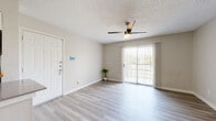 This single-floor unit offers a clean, contemporary design throughout, finished with light-toned luxury vinyl plank flooring that runs seamlessly across all living areas. The open-concept layout connects the entryway directly to the living and dining areas, creating an airy, cohesive flow. A smart thermostat panel is conveniently located near the front door.

The kitchen is thoughtfully designed with white cabinetry, a classic subway tile backsplash, light countertops, a peninsula with additional workspace, and a full suite of stainless steel appliances including a refrigerator and dishwasher. A flush-mount ceiling fixture provides practical illumination in this space.

The living area benefits from abundant natural light streaming through a large sliding glass door fitted with vertical blinds, offering outdoor views and direct access to an exterior area. A ceiling fan enhances comfort throughout the main living space.

The bedroom is generously sized with two windows dressed in horizontal blinds, allowing natural light from multiple directions. A ceiling fan with a light kit provides additional ventilation and ambiance.

The bathroom features a clean, all-white aesthetic with a full soaking tub and tiled shower surround, paired with matching white fixtures. Throughout the unit, textured ceilings, neutral warm-beige walls, and crisp white trim contribute to a bright and cohesive interior atmosphere.