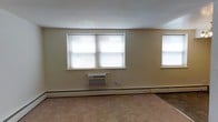 This single-floor space offers a straightforward and functional layout with a clean, neutral aesthetic. The interior is finished with light beige walls that provide a warm, understated backdrop throughout the space. Carpeted flooring adds a layer of comfort underfoot, contributing to a quiet and cozy atmosphere.

Natural light is brought in through multiple windows fitted with horizontal blinds, allowing for adjustable light control and privacy. The windows are positioned to illuminate the interior effectively, giving the space a bright and open feel.

Climate comfort is addressed through a combination of a wall-mounted window air conditioning unit and baseboard heating that runs along the perimeter of the room, ensuring temperature regulation across all seasons. The baseboard heating system is a practical and consistent feature of the space.

The overall design is utilitarian and uncluttered, making the space highly adaptable for a variety of uses. The neutral color palette, practical fixtures, and functional layout offer a solid foundation for personalization or professional use. This is a no-frills space that prioritizes comfort and functionality in equal measure.