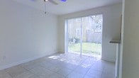This two-story space offers a clean, airy aesthetic throughout, characterized by light neutral walls and well-maintained tile flooring that reflects abundant natural light. The living area is a standout feature, anchored by large sliding glass doors fitted with vertical blinds that open directly to a verdant outdoor space, complete with a patio seating area and a well-kept lawn. Sunlight streams generously through the expansive glass, creating a warm, welcoming atmosphere and a seamless connection between the interior and exterior. A ceiling fan overhead adds both functional comfort and a casual touch to the room. The layout spans two floors, offering a practical separation of living spaces that suits a variety of lifestyles. The overall ambiance of the space is bright and open, with a simple, uncluttered design that allows each room's architectural details — from the tile work underfoot to the generous glazing — to take center stage. The outdoor views visible from the main living area lend the space a refreshing, nature-connected feel throughout the day.