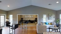 Located in Arlington, Texas, this single-story space makes a strong first impression with its dramatic vaulted ceiling that rises to a sharp peak, creating a sense of openness and grandeur throughout the main area. The interior is finished in a soft, neutral gray palette that lends a calm and professional ambiance.

The main open area features warm hardwood flooring and is furnished with a mix of casual and functional seating, including a patterned lounge chair accented with teal throw pillows and a sleek bistro-style high-top table with modern bar stools — ideal for relaxed gatherings or informal meetings.

Adjacent to the lounge area, a dedicated workstation alcove is recessed into the space, offering multiple computer workstations set along a wide desk surface. The alcove is well-appointed with framed artwork on the walls, adding a polished, finished look to the work environment. Natural light filters in through a window on the side wall, brightening the space and connecting it to the outdoors.

Overall, this space blends comfort and functionality seamlessly, making it well-suited for community use, collaborative work, or quiet productivity in a thoughtfully designed, inviting environment.