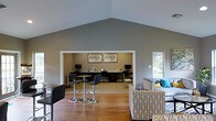 Located in Arlington, Texas, this single-story space makes a strong first impression with its dramatic vaulted ceiling that rises to a sharp peak, creating a sense of openness and grandeur throughout the main area. The interior is finished in a soft, neutral gray palette that lends a calm and professional ambiance.

The main open area features warm hardwood flooring and is furnished with a mix of casual and functional seating, including a patterned lounge chair accented with teal throw pillows and a sleek bistro-style high-top table with modern bar stools — ideal for relaxed gatherings or informal meetings.

Adjacent to the lounge area, a dedicated workstation alcove is recessed into the space, offering multiple computer workstations set along a wide desk surface. The alcove is well-appointed with framed artwork on the walls, adding a polished, finished look to the work environment. Natural light filters in through a window on the side wall, brightening the space and connecting it to the outdoors.

Overall, this space blends comfort and functionality seamlessly, making it well-suited for community use, collaborative work, or quiet productivity in a thoughtfully designed, inviting environment.