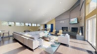 This single-floor communal space is defined by its striking architectural character and thoughtfully designed gathering areas. The centerpiece of the main lounge is a soaring vaulted ceiling that rises dramatically above a large-scale, gray-tiled dual fireplace wall, which anchors the room with a bold, modern aesthetic. A wall-mounted television is positioned above the fireplace, making it a true focal point for socializing and relaxation.

The living area is generously proportioned, accommodating a large sectional sofa and additional seating chairs arranged around a geometric-patterned area rug and a sleek wood coffee table. Light wood-tone flooring runs throughout, complementing the warm yellow accent walls and neutral gray tones. Natural light pours in through a row of clerestory windows along one wall and a tall arched window arrangement, providing bright, open views of the surrounding greenery.

A casual dining nook, framed by navy blue drape curtains and a window overlooking lush trees, offers an intimate seating option with a round table and mixed chairs. Adjacent to this is a compact kitchenette outfitted with dark cabinetry, a stainless steel refrigerator, and a bar-height counter with stylish bar stools beneath a trio of gold globe pendant lights — ideal for casual entertaining or refreshments.