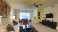 This single-story space centers around a well-appointed living area that combines classic architectural details with a comfortable, open layout. Decorative white columns frame the room, adding a touch of elegance and helping to define separate zones within the open floor plan. The large picture window along the far wall allows abundant natural light to flood the interior, creating a bright and welcoming atmosphere. Neutral, light-toned walls complement the carpeted flooring throughout, contributing to a warm and cohesive aesthetic. A ceiling fan with a brushed nickel finish is centrally positioned overhead, providing both function and a subtle design element. The living area comfortably accommodates a white sofa, an accent armchair in warm terracotta tones, and a dark-finished rectangular coffee table anchored by a boldly patterned area rug in brown and black tones. A console or sideboard near the entryway zone offers additional storage and display space, while framed wall art adds visual interest. A dedicated sitting or writing area is positioned near the window, making excellent use of the natural light. Overall, the space presents a balanced blend of classic detailing and relaxed comfort.