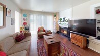 This single-story residence is thoughtfully laid out across a compact yet functional floor plan. The living room serves as the heart of the home, featuring a fireplace flanked by a wall-mounted television, wood-look flooring, and a large sliding glass door fitted with vertical blinds that invite natural light and provide outdoor access. The space flows openly into a defined dining and office area, creating a versatile zone for both work and daily living.

The galley-style kitchen is efficiently designed with white cabinetry, light countertops, and a full suite of appliances including a refrigerator, dishwasher, and range. A doorway at the end of the kitchen connects to additional areas of the unit.

The two bedrooms are carpeted and comfortably sized. One features closet access and an open layout, while the other includes a window with blinds and wall-mounted shelving for display and storage.

The bathroom is a standout feature, stretching as a long corridor-style space with wood-look flooring, an extended vanity with ample cabinetry and counter space, and a separate water closet housing the toilet and shower behind a curtain. Neutral tones and clean finishes carry throughout, contributing to a cohesive and practical ambiance.