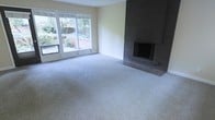 This single-floor space offers a clean, understated layout designed for comfortable living. The main living area is generously proportioned, finished with plush, light-toned wall-to-wall carpeting that adds warmth and softness underfoot. A striking floor-to-ceiling brick fireplace anchors one wall, clad in dark grey tile that extends to a raised hearth, serving as a natural focal point for the room. Opposite the fireplace, a wide set of sliding glass doors and accompanying windows flood the space with abundant natural light, while framing a verdant outdoor view of mature landscaping and a patio area beyond. The light, neutral wall tones complement the soft carpeting and create a calm, airy ambiance throughout. The overall design is practical and timeless, with an open feel that lends itself well to a variety of furniture arrangements. The seamless connection between the interior living space and the outdoor area through the expansive glass doors enhances the sense of openness, making the space feel bright and connected to its natural surroundings.