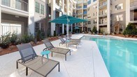 This outdoor amenity space is centered around a generously sized swimming pool with crystal-clear blue water, complemented by a separate spa or hot tub nestled along the pool's edge. The pool deck is finished in a light-toned concrete surface, providing ample room for lounging and circulation. Scattered throughout the deck are mesh sling-style chaise lounges paired with side tables and teal accent umbrellas, creating a cohesive and inviting outdoor retreat.

The courtyard is enclosed on multiple sides by a contemporary multi-story building featuring a neutral gray and beige facade with large windows, black-railed private balconies, and vertical column accents. The architectural design creates a semi-private, resort-like atmosphere. Ground-level access points with glass doors connect the pool area directly to interior common spaces.

Landscaping along the perimeter includes neatly trimmed evergreen shrubs, cypress-style trees, and mulched planting beds that provide greenery and visual separation from the building's base. An outdoor dining and grilling area with seating is visible in the background, adding functional versatility to the space. The open courtyard orientation allows for abundant natural daylight throughout the day, enhancing the overall ambiance of this well-appointed amenity area.