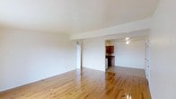 This single-floor space is defined by its warm, light-toned hardwood floors that extend throughout the open living area, lending a natural, inviting character to the interior. The main living space is generously proportioned, with smooth white walls and a white ceiling that enhance the sense of openness and airiness. A structural beam is subtly integrated into the ceiling, adding a modest architectural detail without disrupting the clean aesthetic.

The living area flows openly toward the kitchen, which is partially visible from the main room. The kitchen features rich, dark wood cabinetry that provides a warm contrast to the lighter tones of the surrounding space. A ceiling fan with a light fixture is mounted above the kitchen area, supporting both ventilation and ambient lighting. White appliances are also visible, contributing to a classic, functional kitchen setup.

A white interior door is accessible from the living area, suggesting a connected hallway or additional room. The overall design is simple and well-maintained, with a neutral color palette that creates a versatile and adaptable living environment throughout the single-floor layout.