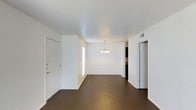 This single-floor space offers a clean, open layout that seamlessly connects the living and dining areas, creating a sense of spaciousness and flow throughout. The interior is defined by rich, dark hardwood flooring that runs continuously across the main living areas, providing a warm and grounded aesthetic that contrasts beautifully with the crisp white walls and trim. The textured popcorn ceiling adds a classic touch overhead, while a delicate multi-arm chandelier serves as a focal point in the dining area, adding a refined decorative element to the space. Natural light filters in through a window near the front entry, brightening the interior and enhancing the open feel. The front entry features a solid paneled door with a bright white finish, providing a welcoming arrival point. A hallway extends toward additional rooms at the rear, hinting at further functional spaces beyond the main living zone. Visible HVAC vents are integrated into the ceiling, ensuring year-round climate comfort. The overall ambiance is bright, neutral, and move-in ready, offering a versatile canvas well-suited for a variety of interior design styles and furnishing preferences.