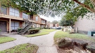 This single-floor residence is situated within a well-established multi-unit complex characterized by warm brick and wood-panel exteriors, metal stairways, and a landscaped courtyard with mature trees, green lawns, and a winding concrete walkway that adds to the property's welcoming curb appeal.

Inside, the interior reflects a thoughtfully curated aesthetic. The dining area serves as a standout feature, anchored by a striking round green marble pedestal table accompanied by woven rattan chairs and set atop a textured area rug. A large-scale framed artwork in neutral tones adorns the wall, contributing to the sophisticated atmosphere of the space. Recessed lighting illuminates the room from above, while dark stained concrete floors ground the design with an industrial-chic sensibility.

Distinctive architectural details include a vintage-style set of double doors with aged patina finishes that open to an adjoining room, as well as a sleek sliding barn door on a black rail track, offering both functional separation and visual interest. The neutral white walls throughout provide a clean, gallery-like backdrop that allows the carefully selected furnishings and artwork to take center stage. The overall ambiance is one of refined eclecticism, blending raw materials with curated décor for a unique and characterful living environment.
