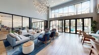 This contemporary community clubhouse is designed to deliver an upscale lifestyle experience through thoughtfully curated amenity spaces. The building's exterior reflects a modern multi-story residential complex, with a central courtyard pool deck framed by textured stone walls, glass panel railings, and native plantings including tall succulents and a living green wall feature.

Inside, the main lobby commands immediate attention with soaring double-height ceilings and a dramatic cascading chandelier of elongated brass-tipped pendants. Floor-to-ceiling glass doors connect seamlessly to the outdoor pool terrace, flooding the interior with abundant natural light. The open lounge area features a curated mix of neutral and teal upholstered seating arranged on area rugs over light wood-look tile flooring.

Adjacent spaces include a social lounge with teal banquette seating, a wall-mounted television, and a shuffleboard table, as well as intimate booth-style nooks with channel-tufted seating and curated artwork. A wet bar area showcases a striking large-format marble slab backsplash, concrete countertops, and striped bar stools. Glass-panel doors throughout maintain visual connectivity between zones, while a fitness area is accessible via black-framed glass doors, rounding out this well-appointed amenity level.