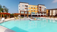 Located in Seneca, South Carolina, this outdoor amenity space is designed to deliver a resort-style experience for its residents. At the heart of the area is a generously sized, crystal-clear swimming pool with a stamped concrete surround that adds a refined, textured aesthetic to the deck. The pool deck is outfitted with rows of lounging chairs, providing ample space for sunbathing and relaxation under the South Carolina sky.

A striking wooden pergola anchors one end of the space, paired with an outdoor stone fireplace that creates a warm, inviting gathering area. Decorative stone masonry columns and paving continue the upscale design language throughout. On the opposite end of the pool, a large outdoor movie screen mounted to a stone-clad wall serves as a standout entertainment feature, ideal for outdoor movie nights by the water.

The surrounding multi-story residential building features a bold contemporary façade, combining yellow and gray paneling with brick accents, large windows, and private balconies that overlook the pool area. Lush mature trees frame the perimeter, providing natural privacy and shade. The entire space is enclosed by a sleek iron fence, maintaining a sense of security while preserving the open, airy ambiance of this well-appointed outdoor retreat.