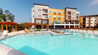 Located in Seneca, South Carolina, this outdoor amenity space is designed to deliver a resort-style experience for its residents. At the heart of the area is a generously sized, crystal-clear swimming pool with a stamped concrete surround that adds a refined, textured aesthetic to the deck. The pool deck is outfitted with rows of lounging chairs, providing ample space for sunbathing and relaxation under the South Carolina sky.

A striking wooden pergola anchors one end of the space, paired with an outdoor stone fireplace that creates a warm, inviting gathering area. Decorative stone masonry columns and paving continue the upscale design language throughout. On the opposite end of the pool, a large outdoor movie screen mounted to a stone-clad wall serves as a standout entertainment feature, ideal for outdoor movie nights by the water.

The surrounding multi-story residential building features a bold contemporary façade, combining yellow and gray paneling with brick accents, large windows, and private balconies that overlook the pool area. Lush mature trees frame the perimeter, providing natural privacy and shade. The entire space is enclosed by a sleek iron fence, maintaining a sense of security while preserving the open, airy ambiance of this well-appointed outdoor retreat.