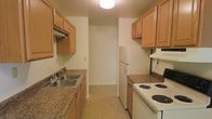This single-floor space centers around a galley-style kitchen that serves as its defining feature. The kitchen is laid out with countertops and cabinetry running along both sides, creating an efficient corridor-style workflow. Warm-toned wooden cabinets line the upper and lower sections of both walls, providing generous storage space throughout. On one side, a double-basin sink sits beneath the cabinetry with a range hood installed above, accompanied by granite-look laminate countertops that extend to the lower cabinets below. The opposite wall houses a white electric coil-top range with an overhead ventilation unit, along with additional counter space and lower cabinetry. A full-size white refrigerator is positioned at the end of one wall. The flooring throughout the kitchen appears to be a neutral-toned resilient tile, practical for high-traffic use. A single overhead light fixture provides general illumination for the space. At the far end of the kitchen, a doorway opens into an adjacent area, suggesting a connected layout beyond the kitchen. The overall design prioritizes functionality and efficient use of available space.