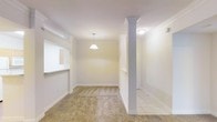 This single-floor residence showcases a thoughtfully designed open-concept layout that connects the living and dining areas in a cohesive, flowing arrangement. Crown molding runs throughout the space, lending a refined architectural touch to the otherwise clean, neutral palette of white walls and beige tones. A distinctive white decorative column serves as a subtle visual divider between the living and dining zones while preserving the open feel of the interior.

The living area is finished with wall-to-wall carpeting in a warm beige tone, providing a soft and comfortable underfoot feel, while the adjacent dining and entryway areas transition to light-toned tile flooring. A pendant light fixture hangs above the dining area, offering focused illumination and a simple, classic design accent.

A pass-through counter or bar-height ledge connects the main living space to what appears to be a kitchen area, making the layout practical for entertaining and everyday living. Recessed and flush-mount ceiling lights provide additional illumination throughout the space. The overall ambiance is bright, neutral, and move-in ready, with architectural details that elevate the standard finishes into a polished, cohesive living environment.