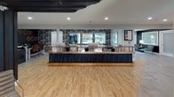 This thoughtfully designed single-floor commercial space combines functionality with contemporary style across several distinct zones. The centerpiece of the layout is a striking reception or service counter finished in warm wood tones with a navy blue paneled front accented by teal linear details. Behind it, a bold black-and-white patterned accent wall adds visual energy, while large windows flood the area with natural light and offer views of the surrounding exterior.

Adjacent to the main counter area, a casual seating zone features round live-edge wooden tables paired with modern metal chairs in black and teal. Copper geometric pendant lights hang overhead, and a series of vibrant blue and teal geometric artworks line the gray-painted walls, reinforcing a cohesive color palette throughout.

A separate nook near a large picture window provides additional seating with outdoor views, creating a bright and welcoming atmosphere. Toward the rear of the space, a compact kitchenette is outfitted with navy and teal cabinetry, open wood shelving, and a small appliance station, conveniently positioned near a glass-paneled exit door. Light wood-look flooring and large-format tile flooring transition seamlessly between zones, unifying the space with a clean, modern aesthetic.