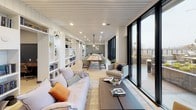 This single-floor workspace is designed to support a variety of professional and collaborative needs within a cohesive, well-appointed environment. The layout, as revealed in the floor plan, organizes distinct functional zones that flow naturally into one another while maintaining visual consistency throughout.

The central lounge area serves as the social heart of the space, furnished with plush sofas, a coffee table, and a patterned area rug atop light hardwood flooring. Built-in white shelving lines the walls, filled with books and décor, while a slatted white ceiling with recessed lighting adds architectural texture. Floor-to-ceiling black-framed glass doors open to an outdoor terrace, filling the space with natural light and views of the exterior.

Adjacent to the lounge is a communal dining area featuring a long wooden table surrounded by upholstered chairs, anchored beneath a pendant light. The conference room is distinguished by deep slate-blue wainscoting, a rustic wood table, leather chairs, a wall-mounted display screen, and a modern globe chandelier. Framed black-and-white architectural photographs accent the walls throughout, lending the space a curated character.

A compact workstation area provides two dedicated computer desks set beneath paired industrial-style sconce lighting, offering a focused setting for individual tasks.