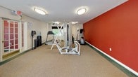 This fitness center is a single-level facility thoughtfully designed to accommodate a diverse range of exercise needs. The space is divided into distinct functional zones, each outfitted with purpose-driven equipment. The main area features a multi-station cable weight machine as a central anchor, complemented by treadmills, an elliptical trainer, and stationary and recumbent bikes for cardiovascular training. A dedicated stretching and floor exercise zone includes yoga mats, stability balls, and an assortment of dumbbells and resistance accessories.

The interior design incorporates bold red accent walls that energize the space, contrasting with neutral beige carpeted flooring and white walls. Ceiling fans and flush-mounted light fixtures provide comfortable ventilation and consistent illumination throughout. A full-length mirrored wall along one side visually expands the room while allowing users to monitor their form. Natural light enters through a window dressed with dark curtains, offering a pleasant connection to the surrounding greenery. A wall-mounted flat-screen television adds an entertainment option for cardio sessions. The space is well-organized, practical, and equipped to support a comprehensive fitness routine.