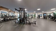 This sophisticated fitness center is designed to deliver a comprehensive workout experience across multiple well-defined zones. The main floor is finished with durable dark rubber flooring and features an extensive array of strength and cardio equipment, including multi-station cable machines, free weights, and weight benches, all arranged to maximize space and ease of movement. Circular recessed lighting illuminates the space evenly throughout.

A dedicated cardio zone lines one wall beneath a warm wood-paneled ceiling accent, housing a full row of treadmills and elliptical machines, each equipped with individual digital displays. Large mirrors expand the sense of space and allow users to monitor their form during workouts.

A separate studio room, accessible through sleek floor-to-ceiling glass and black-framed partition doors, features light wood-look flooring and is equipped with spin bikes, rowing machines, a power rack with boxing bags, and suspension training equipment. A striking illuminated living green wall with an X-shaped design adds a vibrant, biophilic focal point to the studio.

The main floor also incorporates a free-weight area with organized dumbbell racks, multiple cable and resistance machines, and wall-mounted televisions. The overall aesthetic is contemporary and polished, blending functionality with upscale design to create an inviting, high-performance fitness environment.