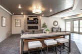 Located in Clear Lake, Iowa, this generously sized single-floor community living room offers a well-appointed, multi-functional gathering space designed for comfort and recreation. The open layout is anchored by light-toned hardwood-style flooring that flows throughout the main area, complemented by a neutral, contemporary color palette.

A standout feature is the dedicated lounge zone, which showcases a shiplap-style ceiling with recessed and pendant lighting, a built-in fireplace framed by floor-to-ceiling tile, and a wall-mounted television. Comfortable swivel chairs and a sofa surround a wooden coffee table, creating an inviting retreat within the larger space.

The recreation area takes center stage with a full-size pool table, while a high-top bar counter with bar stools provides additional social seating. A dining area with a round table and modern chairs adds further versatility to the layout.

Large glass double doors and flanking windows along one wall flood the space with natural light and provide direct views and access to the outdoors. Recessed lighting throughout ensures the interior remains bright and well-lit. The overall design blends modern and transitional elements, making this community space equally suited for relaxed gatherings and active recreation.