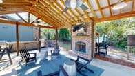 This thoughtfully designed outdoor living space in Aiken, South Carolina offers a welcoming retreat built for year-round enjoyment. The centerpiece of the pavilion is a full-height brick wood-burning fireplace, complete with an arched firebox opening and a wall-mounted television above the mantel, creating an ideal gathering focal point. The structure is defined by an impressive vaulted ceiling of exposed wood beams and rafters, lending a warm, rustic character to the open-air environment. Multiple ceiling fans with industrial-style pendant lights are suspended throughout, ensuring comfort even during warm South Carolina days. The spacious concrete floor is anchored by a large area rug and furnished with Adirondack chairs and barrel-style side tables, offering relaxed and inviting seating around the fireplace. A dedicated outdoor kitchen and grilling station with a gas grill and stone surround sits adjacent to the covered area, perfect for entertaining. High-top bar seating provides additional dining options. The open sides of the pavilion frame lush green surroundings and mature trees, filling the space with natural light and garden views. A community pool with iron fencing is visible nearby, accessible via a landscaped walkway, rounding out this exceptional outdoor living destination.