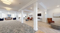 Located at 28 Everett Road, this generously scaled single-floor amenity space offers a sophisticated and welcoming environment designed for comfort and community. The entrance opens onto a striking foyer featuring a herringbone-patterned light wood floor, decorative white columns with crown molding details, and a curated seating area with leather accent chairs and a wall-mounted display screen.

The expansive open living area continues the refined aesthetic with coffered ceilings, additional white columns, and wide-plank light hardwood flooring. Plush seating arrangements in leather and navy blue anchor the space, creating distinct lounging zones that flow naturally into one another.

The combined dining, kitchen, and living area is a standout feature, boasting a stone fireplace, pendant lighting, large windows with exterior views, a communal dining table, and a stylish bar counter with gold-accented glass shelving. The full kitchen is outfitted with white shaker cabinetry, a subway tile backsplash, stainless steel appliances, and light wood flooring.

A salon room enclosed by sleek black-framed glass doors provides a private, professional-grade beauty space with round mirrors and styling chairs. The exercise room rounds out this thoughtfully appointed space, making 28 Everett Road a comprehensive and elegantly designed community destination.