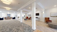 Located at 28 Everett Road, this generously scaled single-floor amenity space offers a sophisticated and welcoming environment designed for comfort and community. The entrance opens onto a striking foyer featuring a herringbone-patterned light wood floor, decorative white columns with crown molding details, and a curated seating area with leather accent chairs and a wall-mounted display screen.

The expansive open living area continues the refined aesthetic with coffered ceilings, additional white columns, and wide-plank light hardwood flooring. Plush seating arrangements in leather and navy blue anchor the space, creating distinct lounging zones that flow naturally into one another.

The combined dining, kitchen, and living area is a standout feature, boasting a stone fireplace, pendant lighting, large windows with exterior views, a communal dining table, and a stylish bar counter with gold-accented glass shelving. The full kitchen is outfitted with white shaker cabinetry, a subway tile backsplash, stainless steel appliances, and light wood flooring.

A salon room enclosed by sleek black-framed glass doors provides a private, professional-grade beauty space with round mirrors and styling chairs. The exercise room rounds out this thoughtfully appointed space, making 28 Everett Road a comprehensive and elegantly designed community destination.