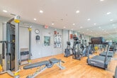Located at 472 Brock Avenue in Burlington, Ontario, this fitness centre is a well-appointed, single-room facility designed to support a comprehensive workout experience. The space features warm hardwood flooring throughout, neutral grey walls, and a bright, recessed lighting system that evenly illuminates the entire room. Large windows fitted with roller blinds allow generous natural light to filter in while offering views of the surrounding area, contributing to an open and energizing atmosphere.

The fitness centre is equipped with a diverse range of commercial-grade exercise equipment. Cardio offerings include treadmills and elliptical trainers from SportsArt, as well as a stationary bike, ensuring varied aerobic training options. A dedicated strength training area features a tiered dumbbell rack stocked with a full range of free weights, along with Hoist-branded weight benches and cable machines for resistance training. A flat-screen television is wall-mounted for entertainment during workouts.

Full-length mirrors line one wall, visually expanding the space and allowing users to monitor their form. The room is finished with a posted fitness centre rules board and motivational fitness photography, reinforcing a professional gym environment. The space is accessed through a secure door and is outfitted with standard safety features including an emergency exit sign and fire alarm system.