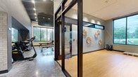 Located in Minneapolis, MN, this well-appointed fitness and wellness facility offers a thoughtfully designed, single-floor layout that balances function and aesthetics. The main workout area features polished concrete flooring, a dark panel ceiling with recessed lighting, and an array of cardio equipment including treadmills, ellipticals, and rowing machines. Full-length mirrors line the walls, while large windows flood the space with natural light and provide views of the surrounding greenery. A dedicated free weights zone includes a dumbbell rack and weight bench, supported by additional mirrored wall sections. Separated by sleek floor-to-ceiling glass partitions with dark bronze framing, a distinct studio area features warm hardwood-style flooring and a full mirrored wall, making it ideal for yoga, stretching, or group fitness. Decorative sunburst wall sculptures add an artistic focal point to this room. A connecting corridor showcases white subway tile accents, open shelving, a water fountain, and a globe pendant light, contributing to the polished, upscale ambiance. Recessed lighting throughout ensures consistent illumination, while the transparent glass partitions maintain a sense of openness and visual continuity across the entire facility.