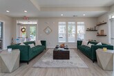 This single-floor space is thoughtfully designed to blend functionality with a welcoming, contemporary aesthetic. Light hardwood-style flooring runs throughout, complementing the neutral white walls and recessed lighting that keep the interior bright and airy.

The main lounge area serves as the heart of the space, featuring plush emerald green sofas, geometric side tables, and a wood coffee table arranged over a soft area rug. Abundant natural light pours in through large windows with white shutters, while French doors provide direct access to the outdoors, enhancing the open, connected feel.

Adjacent to the lounge, a private office area is outfitted with a round conference-style desk and cushioned seating, ideal for one-on-one meetings or consultations. French doors with glass panes allow light to flow freely between areas while maintaining a sense of privacy.

The coffee bar and kitchenette area is a standout feature, showcasing rich dark wood cabinetry with gold hardware, white countertops, and a bold teal accent wall. Wall-mounted screens, a compact refrigerator, and a dedicated coffee station make this a fully functional refreshment hub. Bar-height seating along the counter adds a casual, social element to the space.

Overall, the space exudes a polished yet approachable ambiance, well-suited for a community clubhouse or professional leasing environment.