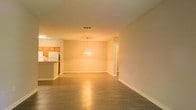 This single-floor space is designed with an open-concept layout that seamlessly integrates the living, dining, and kitchen areas into one cohesive environment. The expansive main area is finished with dark hardwood-style flooring that runs throughout, providing a polished and consistent look underfoot. Neutral-toned walls complement the warm ambient lighting, creating an inviting atmosphere throughout the space.

A chandelier serves as a focal point above the dining area, adding a touch of elegance to the otherwise clean and understated design. The kitchen is positioned to one side and features warm wood cabinetry, a white refrigerator, and additional appliances, all accessible via a raised breakfast bar that doubles as a natural divider between the kitchen and the main living space. The bar provides both functional counter space and an informal seating area.

The layout benefits from a notably open flow, with clear sightlines extending across the full length of the space. A hallway visible toward the rear suggests access to additional rooms. Ceiling-mounted ventilation and recessed lighting fixtures contribute to the practical, well-maintained character of the space overall.